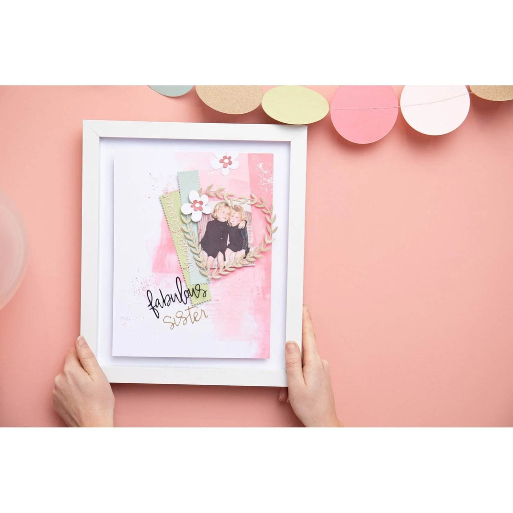 Hände halten einen weißen Bilderrahmen, der eine Scrapbook-Seite mit zwei Personen, dekorativen Papierakzenten und dem Text „Fabulous Sister“ vor einem rosa Hintergrund enthält. Die Kreation zeigt wunderschön das Sizzix Thinlits Die Set Fabulous Frames & Borders von Debi Potter. Oben sind ovale Dekorationen zu sehen, die die Kunst der Kartenherstellung im Scrapbooking demonstrieren.