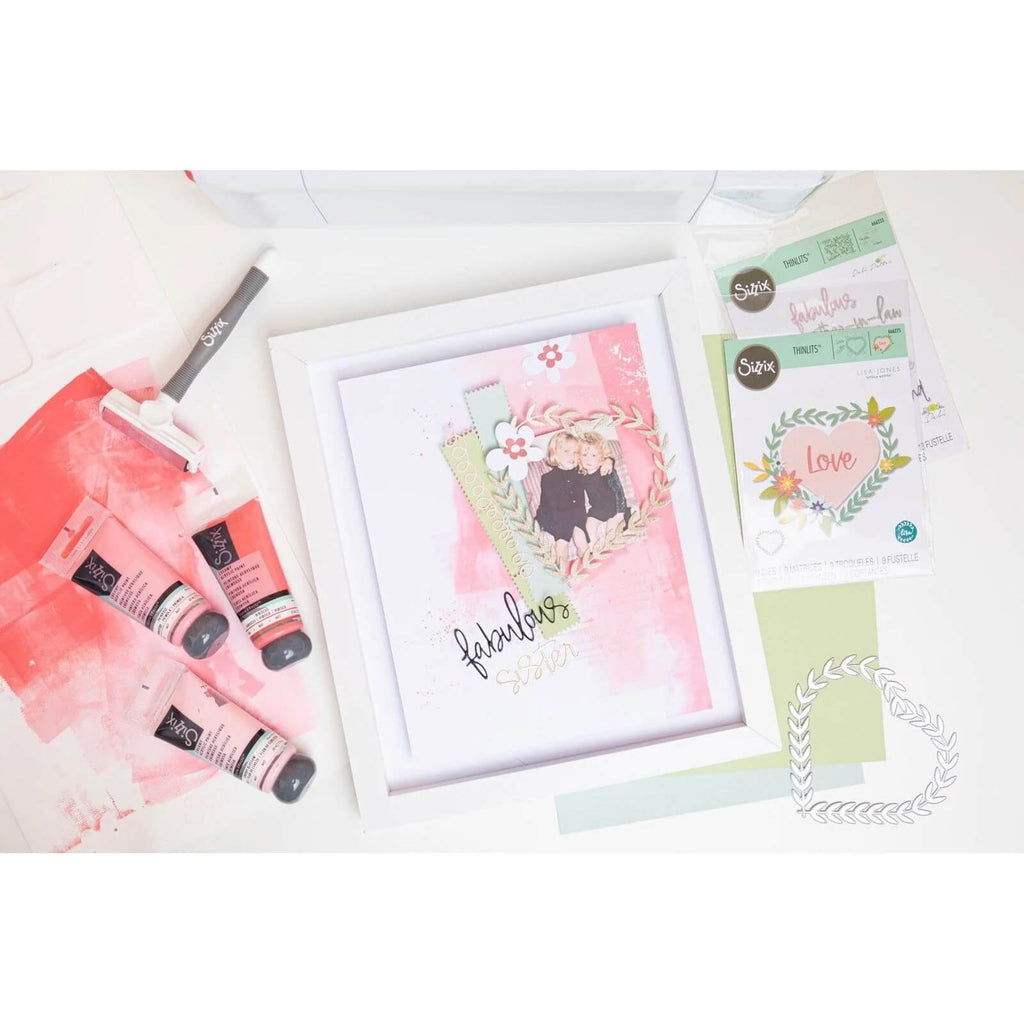 Ein gerahmtes Kunstwerk mit einem Mixed-Media-Design aus Farbtuben, Bastelzubehör und einer hübschen „Love“-Karte darum herum – perfekt für Liebhaber der Kartenherstellung oder des Scrapbooking, alles wunderschön hervorgehoben durch das Sizzix Thinlits Die Set Fabulous Frames & Borders von Debi Potter.