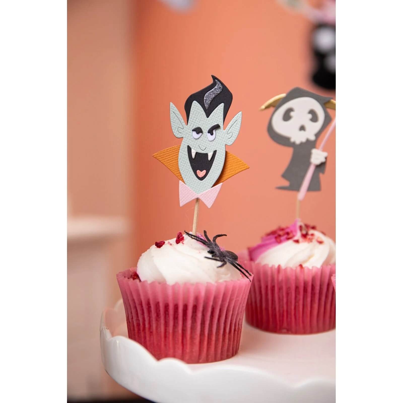 Zwei Cupcakes mit weißem Zuckerguss in rosafarbenen Verpackungen, gekrönt mit Vampir- und Skelett-Papiertoppern sowie einer Plastikspinne - perfekt für Halloween-Papierbastelprojekte mit den Sizzix 666315 - Thinlits mit Stamps Spooky Friends von Sizzix.