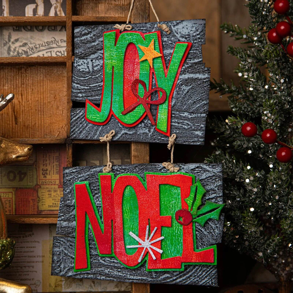 Zwei „Sizzix • Thinlits Stanzschablonen Big Tidings“-Holzschilder mit den Worten „Joy“ und „Noel“ hängen daran, perfekt, um Ihrer Weihnachtsdekoration einen Hauch von Weihnachten zu verleihen.