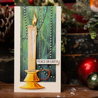 Eine handgemachte Weihnachtskarte mit einer Kerze in einem goldenen Halter, dem Text "PEACE ON EARTH" und grünen Perlengirlanden. Hergestellt mit den Sizzix 666331 Thinlits Stanzschablonen Candleshop Colorize by Sizzix; Ornamente und Tannenzapfen vervollständigen den festlichen Look.