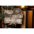 Ein Holzschild, das mit den Sizzix 666337 Thinlits Stanzschablonen Rentierschlitten von Sizzix verziert wurde, trägt die Aufschrift "Santa Stop Here" und befindet sich inmitten von festlicher Weihnachtsdekoration und Büchern.