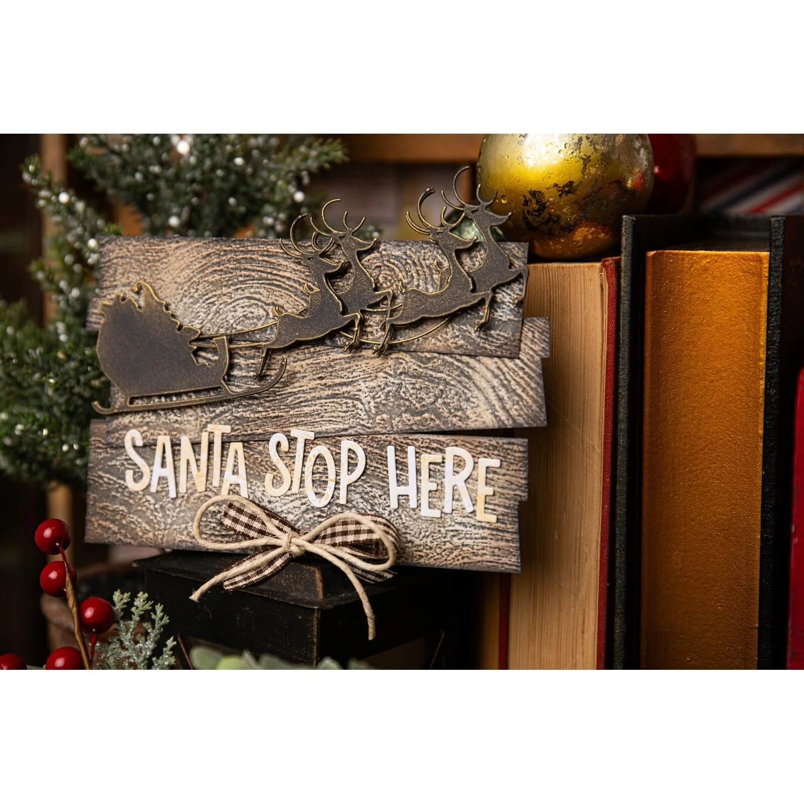 Ein Holzschild, das mit den Sizzix 666337 Thinlits Stanzschablonen Rentierschlitten von Sizzix verziert wurde, trägt die Aufschrift "Santa Stop Here" und befindet sich inmitten von festlicher Weihnachtsdekoration und Büchern.