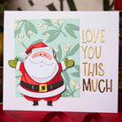 Eine Sizzix 666338 Thinlits Stanzschablonen Weihnachtsgrüße Colorize Karte, von Sizzix, zeigt einen fröhlichen Weihnachtsmann und goldenen "LOVE YOU THIS MUCH" Text auf einem hellgrünen und weißen Hintergrund.