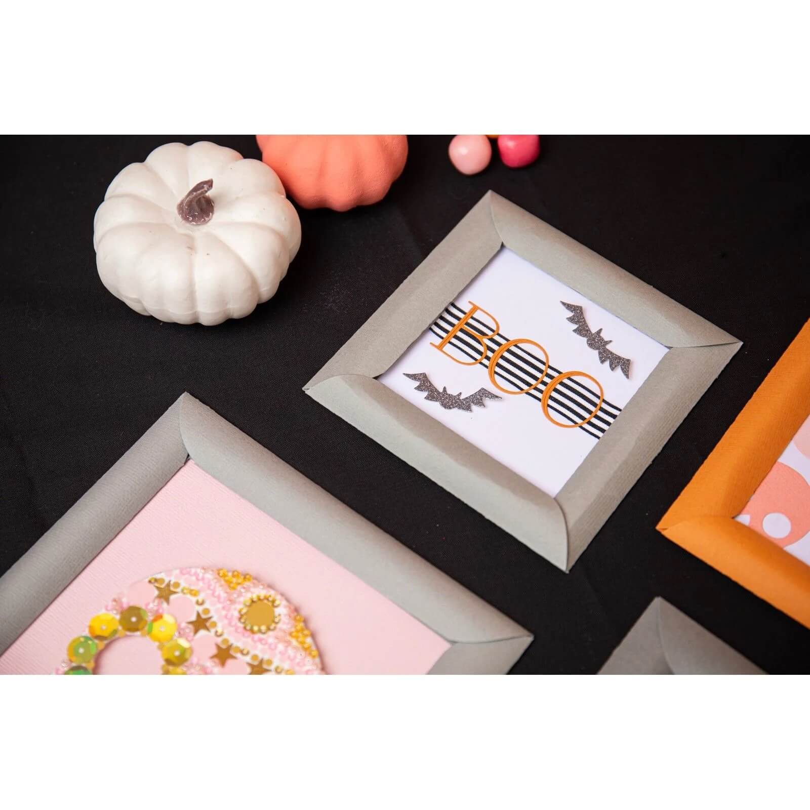 Ein flaches Layout von Halloween-Dekor mit einem weißen Kürbis und "BOO" Fledermauskunst, gestaltet mit Sizzix 666453 - Thinlits Spooky Icons von Sizzix auf einer schwarzen Oberfläche.