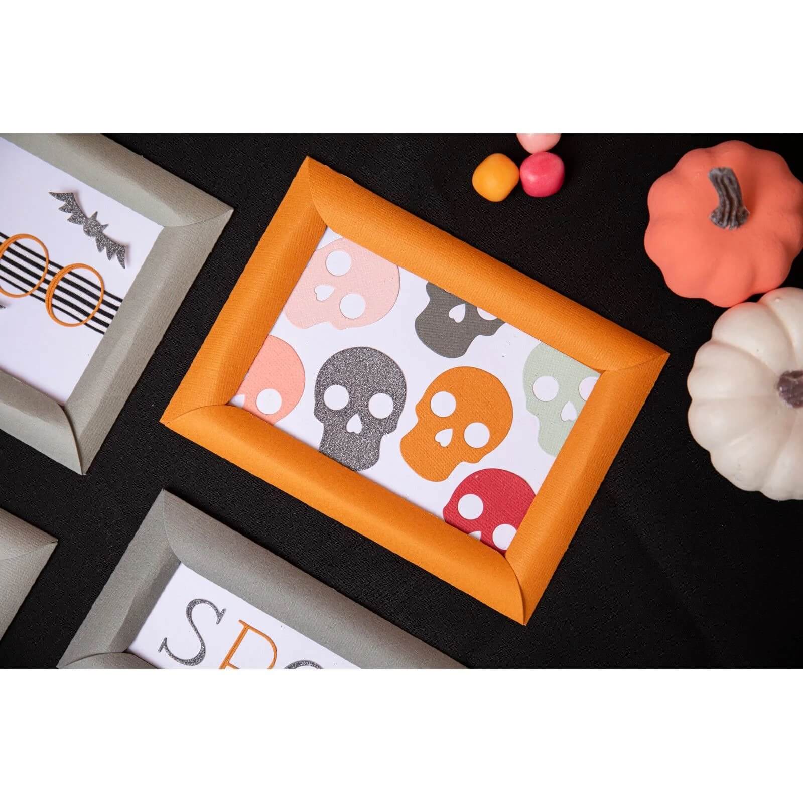 Ein gerahmter Druck mit bunten Totenköpfen liegt auf einer schwarzen Fläche, umgeben von Kürbissen, Halloween-Dekor und Akzenten, die vom Sizzix 666453 - Thinlits Spooky Icons Set von Sizzix inspiriert sind.