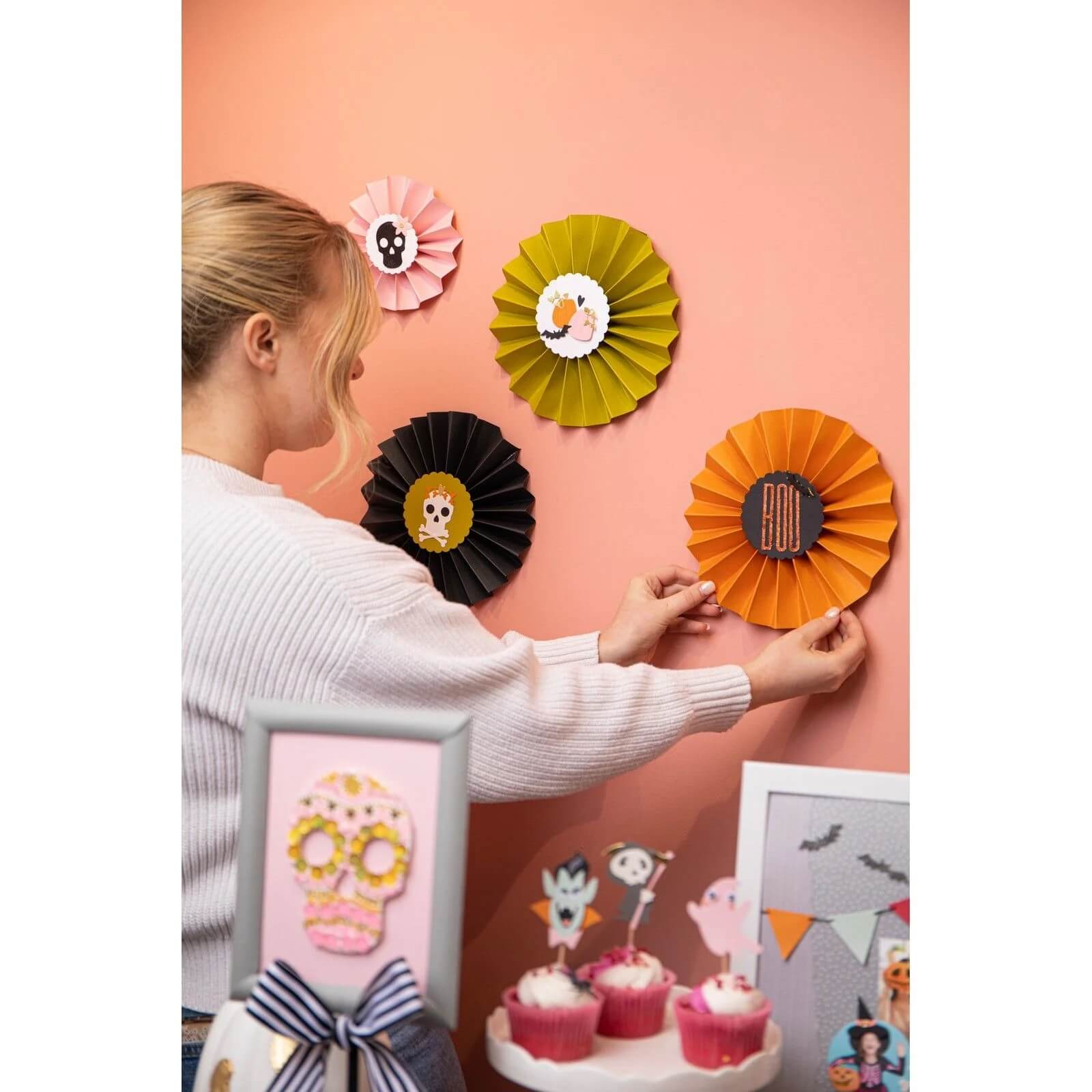 Eine Person dekoriert eine pfirsichfarbene Wand mit Halloween-Papierrosetten, die mit dem Set Sizzix 666453 - Thinlits Spooky Icons von Sizzix hergestellt wurden und über einem Tisch mit Cupcakes und gerahmten Kunstwerken angebracht sind.