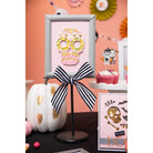 Ein gerahmter Pailletten-Totenkopf auf einem Ständer mit einer schwarz-weiß gestreiften Schleife steht inmitten von Halloween-Dekor - weißer Kürbis, Muffins und Akzente, die mit den Sizzix 666453 - Thinlits Spooky Icons von Sizzix hergestellt wurden.