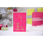 Eine rosafarbene Weihnachtskarte mit den Sizzix Thinlits Variety Sentiments #2 von Sizzix mit dem Schriftzug "Frohe Weihnachten" steht vor verpackten Geschenken und einem weißen Weihnachtsbaum.