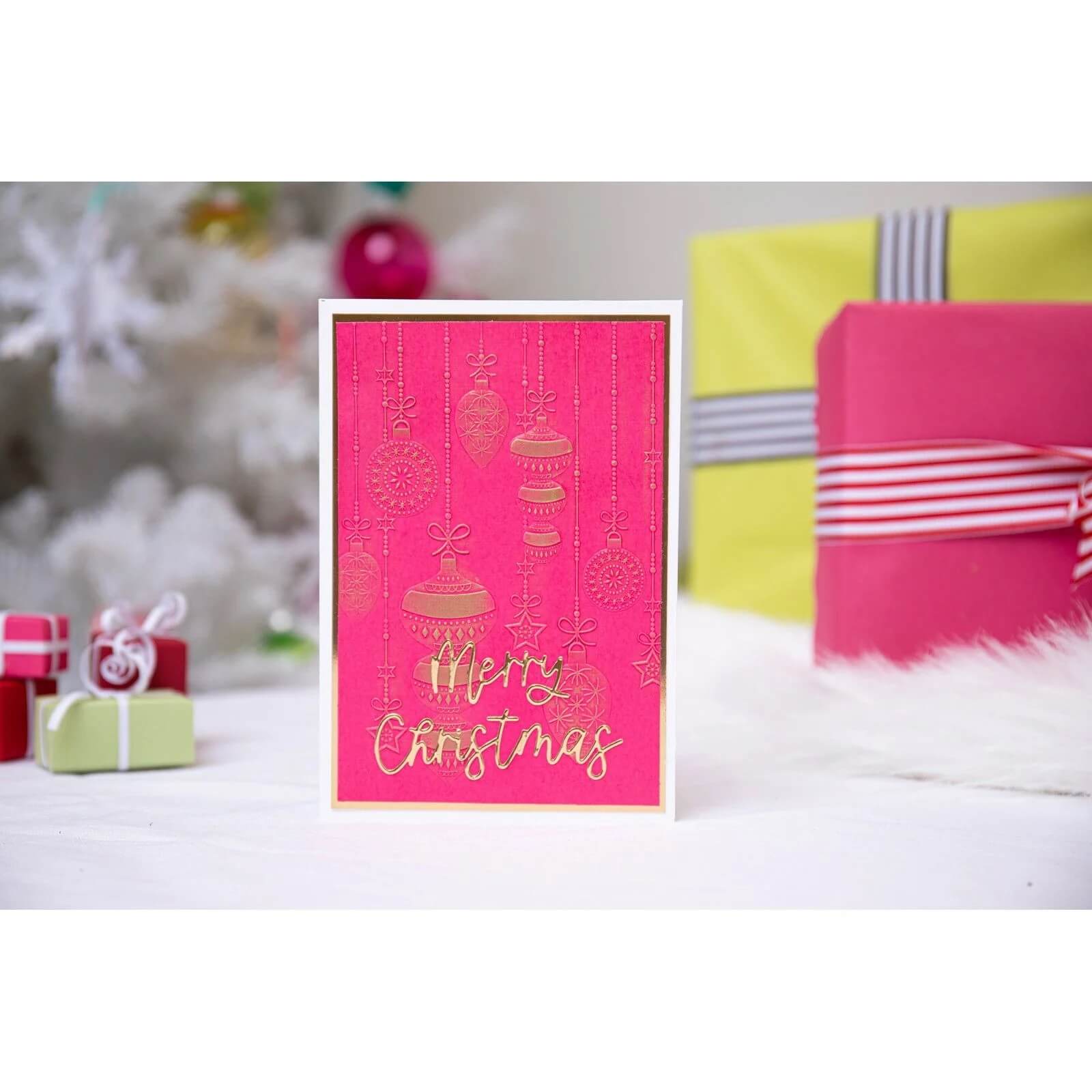 Eine rosafarbene Weihnachtskarte mit den Sizzix Thinlits Variety Sentiments #2 von Sizzix mit dem Schriftzug "Frohe Weihnachten" steht vor verpackten Geschenken und einem weißen Weihnachtsbaum.