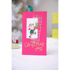 Eine festliche, rosafarbene Weihnachtskarte mit den Sizzix 666457 - Thinlits Variety Sentiments #2 von Sizzix, mit einer Elfe, die ein Geschenk in der Hand hält, karierten Akzenten und dem Schriftzug "Christmas Joy" in Gold - eine charmante Mischung aus Weihnachtsstimmung und kreativem Design.