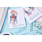 Zwei handgefertigte Karten mit 3D-Hummer- und Krabbenmotiven auf hellblauem Hintergrund, umgeben von Sizzix-Werkzeugen und dem Sizzix 666668 - Clear Stamp Set Seas the Day 11pcs mit charmanten maritimen Sprüchen.