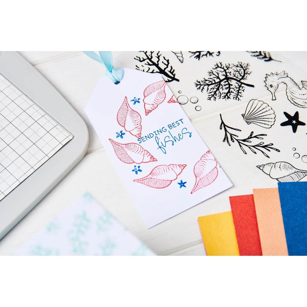 Ein Geschenkanhänger mit dem Text „Sending best fishes“ und Muschelillustrationen, neben einer Schneidematte, Stiften, gemustertem Papier und dem 11-teiligen Sizzix Clear Stamp Set „Seas the Day“ von Sizzix.