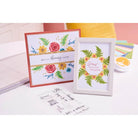 Ein gerahmter Druck und eine Grußkarte mit floralen Motiven, die mit dem Sizzix 666670 Layered Clear Stamp Set Painted Florals (20pcs) von Sizzix hergestellt wurden, liegen auf einem weißen Tisch, umgeben von Bastelzubehör und anderen Bastelmaterialien.