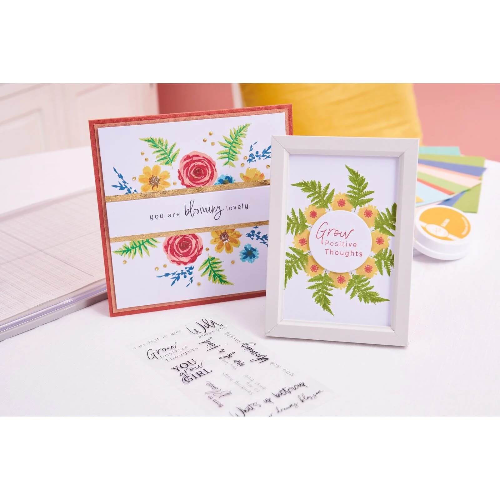 Ein gerahmter Druck und eine Grußkarte mit floralen Motiven, die mit dem Sizzix 666670 Layered Clear Stamp Set Painted Florals (20pcs) von Sizzix hergestellt wurden, liegen auf einem weißen Tisch, umgeben von Bastelzubehör und anderen Bastelmaterialien.