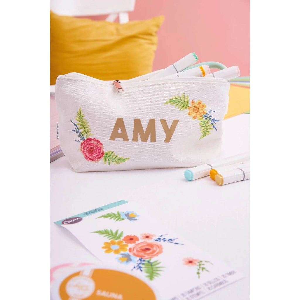 Ein weißer Beutel mit Reißverschluss und der Aufschrift „AMY“ in goldenen Buchstaben, verziert mit Designs aus dem Sizzix Layered Clear Stamp Set Painted Florals (20 Stück), liegt neben bunten Markern und einer Karte mit Blumenmuster auf einem Tisch.