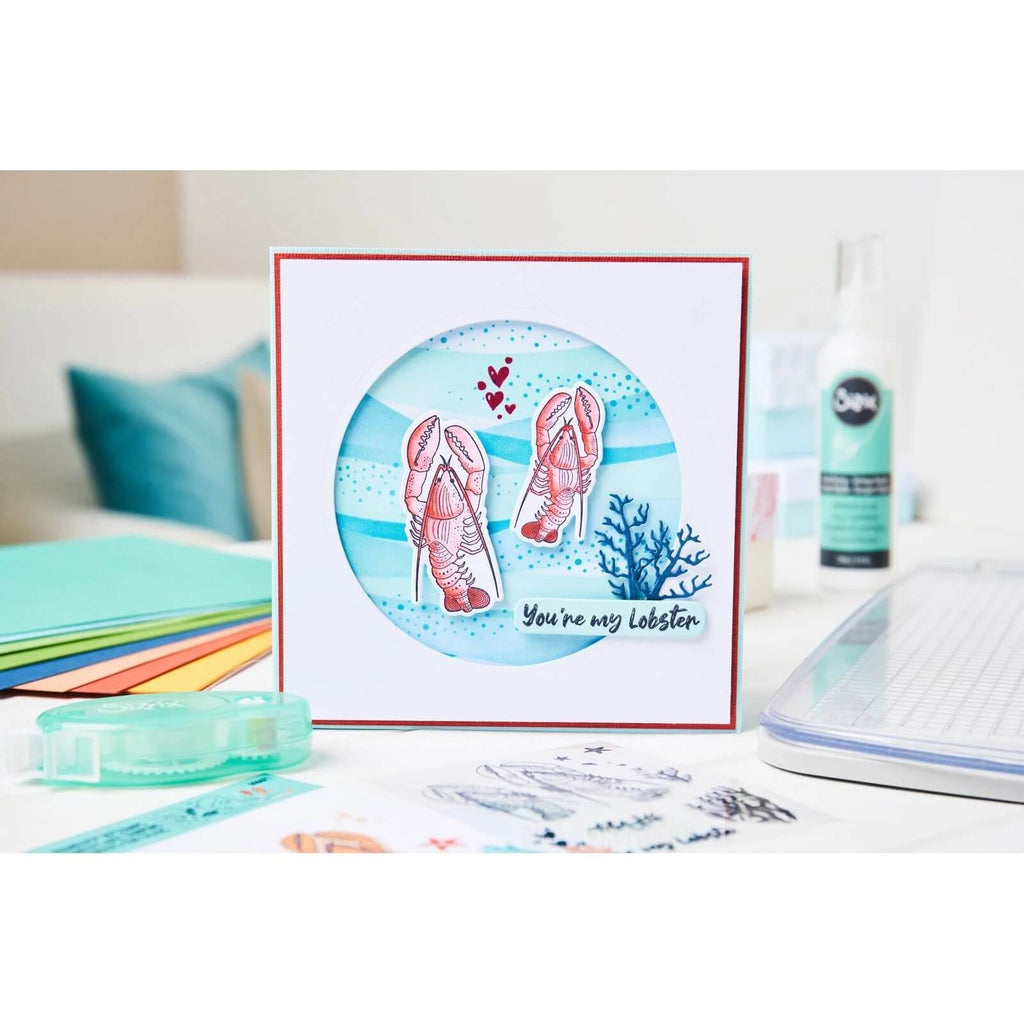 Eine dekorative Karte mit zwei abgebildeten Hummern, kleinen blauen Wellen und dem Text „You're my lobster“ in einem kreisförmigen Ausschnittdesign. Erstellt mit dem Sizzix Framelits Stanzset mit Stempeln „You're my Lobster“ 24-teilig, sind verschiedene Kunstutensilien um die Karte herum auf einem Tisch angeordnet, was das charmante Hummer-Thema noch verstärkt.