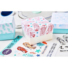 Eine Geschenkschachtel mit Muschel- und Seepferdchenmotiven, die mit dem Sizzix 666669 Clear Stamp Set Ocean Elements (15 Stück) gestempelt wurden, befindet sich zwischen pastellfarbenen Schachteln und Aufklebern mit Meeresmotiven auf einer weißen Fläche.
