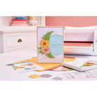 Eine handgefertigte Karte mit geschichteten Papierblumen und Blättern steht auf einem Schreibtisch mit Bastelmaterial, farbigem Papier und einer Sizzix-Schneidemaschine mit dem Sizzix 666672 Thinlits Flowers & Fern 11pcs Set im Hintergrund.