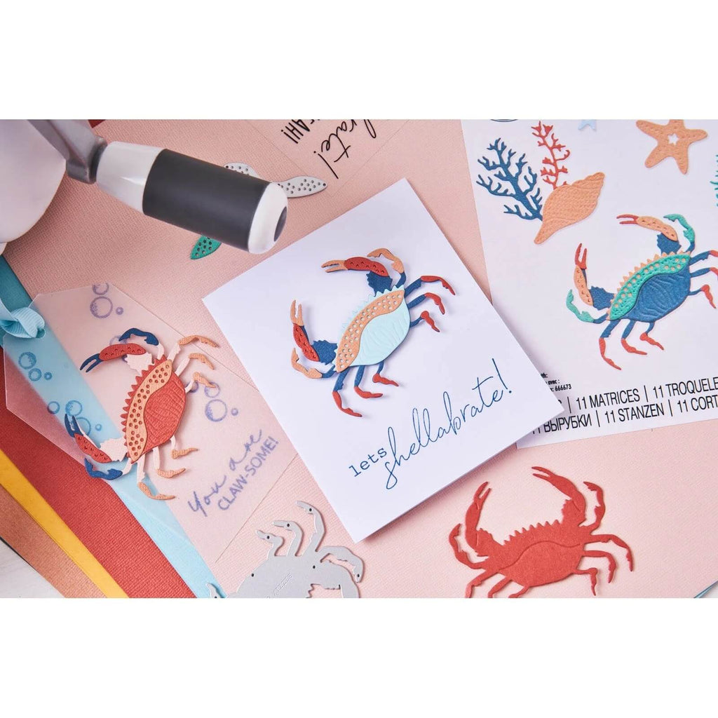 Ein Basteltisch mit Dekorationen zum Thema Krabben, mit einer Karte mit ausgeschnittenen Krabben, die mit dem Sizzix Thinlits Die Set Ocean Critters (10 Stück), farbigem Papier und einem Stempel hergestellt wurden. Auf der Karte steht in leuchtenden Farben „Let's Shellabrate!“. Perfekt für Fans von Hummer-Bastelprojekten!