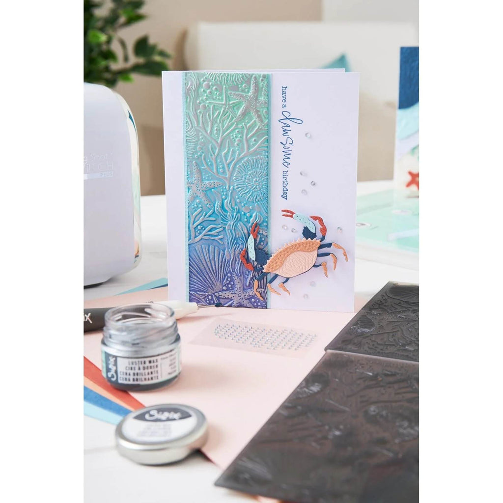 Eine Grußkarte mit einer Unterwasserwelt-Szene mit Koralle und Krabbe liegt auf einem Tisch, neben einem Sizzix • Thinlits Die Set Ocean Medley 11-teilig, Bastelpaste, einem Werkzeug und anderen Bastelmaterialien.