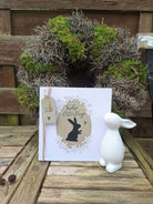 Eine weiße Karte mit schwarzem Kaninchen, ausgestanzt mit der "Stanzschablone: Drei verschiedene Kaninchen" von Stanzenshop.de, steht vor einem Mooskranz - ideal zum Basteln zu Ostern!.