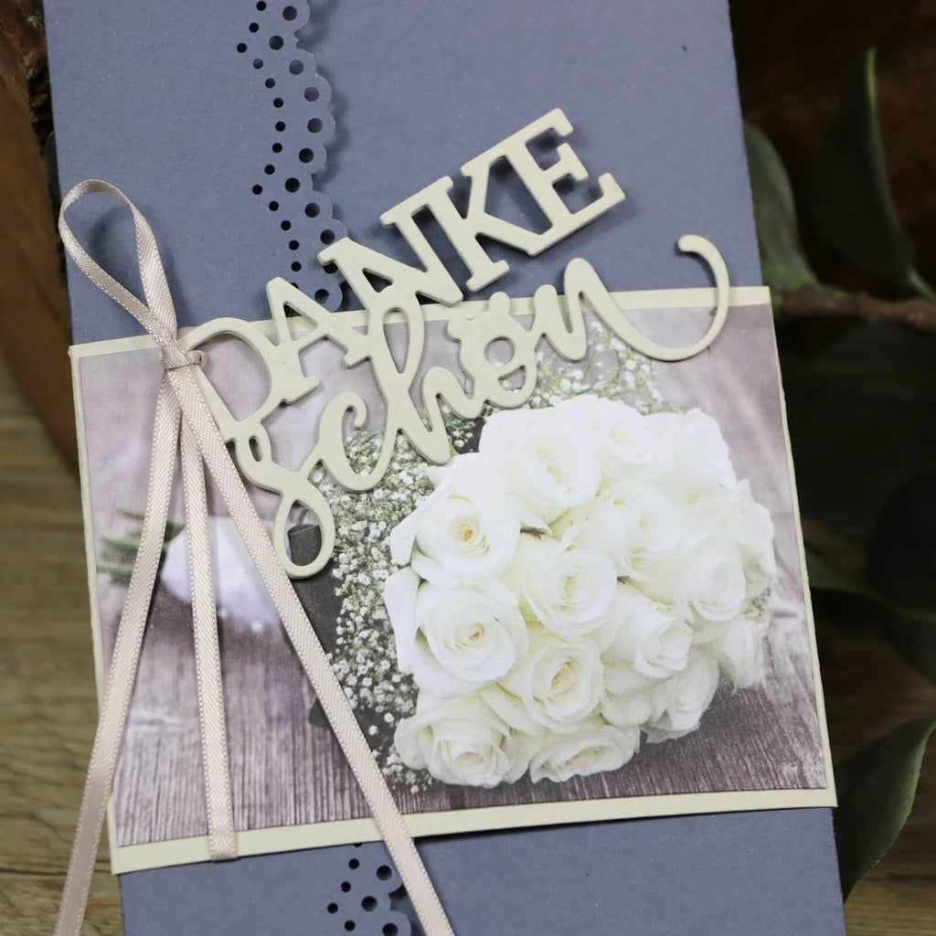 Eine Karte aus der Mundart Stempel-Kollektion von Sizzix mit hellblauem, perforiertem Rand und einem Titelbild mit weißen Rosen. Die 4,1 x 8 cm große Karte ist elegant mit hellbeigen Bändern verziert und trägt auf der Vorderseite die beige Schrift „danke schön“.