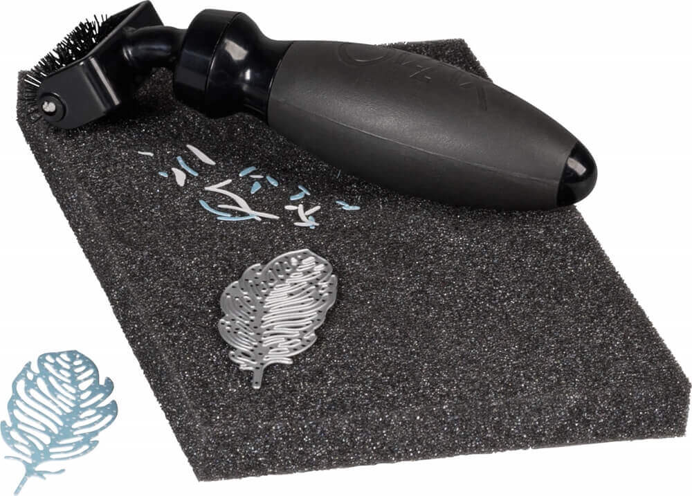 Ein Sizzix Stanz-Zubehör-Set "Die Brush & Foam Pad" (Brush: 14 x 4 cm, Foam Pad: ca. 11,4 x 17,8 cm) in Schwarz ruht auf dem Pad neben einer Metall Thinlits Schablone Federstanze und kleinen Papierstücken.