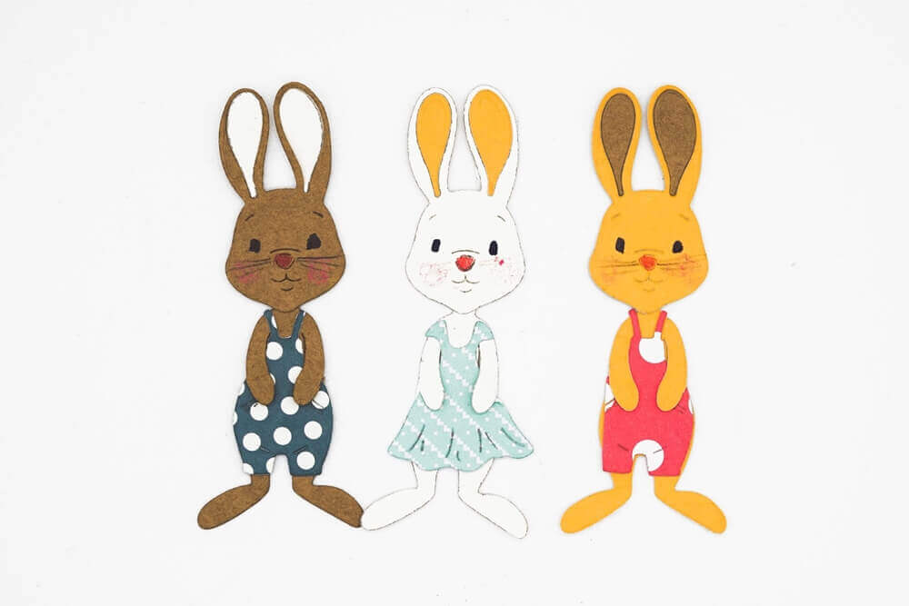 Die Stanzenshop.de Stanzschablone: Bastelset Osterhase zeigt drei Hasenausschnitte in bunten Kleidern, ideal für Scrapbooking oder Basteln. Braun mit marineblauen Punkten, weiß mit blauem Kleid und orange mit rotem Kleid auf weißem Hintergrund.