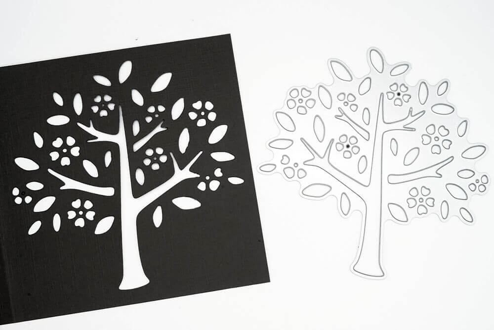 Die Stanzschablone "Baum mit Blüten und Blättern" von Stanzenshop.de liegt neben einem schwarzen Papier-Ausschnitt des Designs - ideal für Bastel- und Scrapbooking-Projekte.