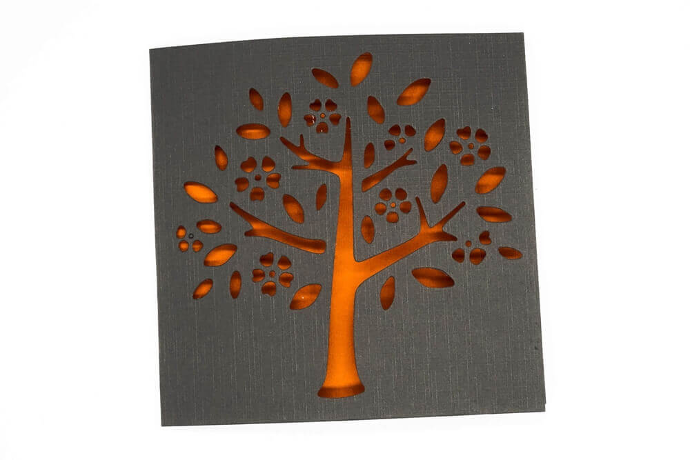 Eine quadratische schwarze Karte mit Baum-, Blüten- und Blattausschnitten leuchtet in orangefarbenem Licht - ideal für Scrapbooking oder Basteln mit der "Stanzschablone Baum mit Blüten und Blättern" von Stanzenshop.de.