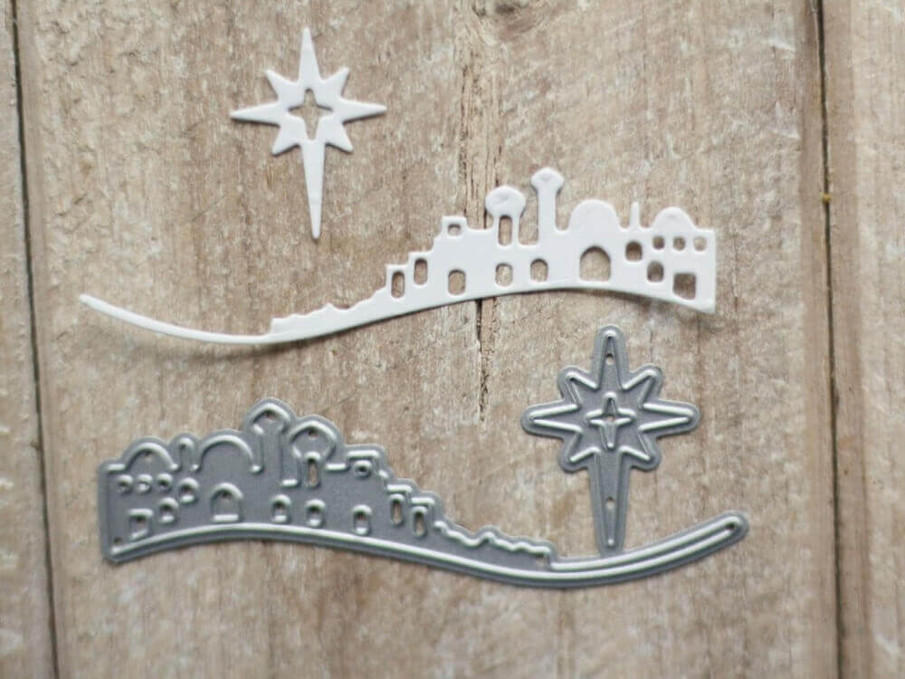 Zwei gestanzte Formen - eine Skyline von Bethlehem und ein Weihnachtsstern, eine weiß und eine metallisch - sind auf Holz abgebildet, ideal für kreative Kartenherstellung oder Basteln im Winter mit Ihrer Stanzschablone: Bethlehem mit Weihnachtsstern von Stanzenshop.de.