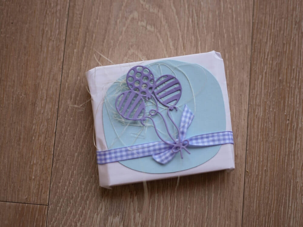 Ein kleines Geschenk, verpackt in weißem Papier mit blauer Schleife und verziert mit der Stanzschablone Drei Luftballons mit Muster von Stanzenshop.de, perfekt zum Basteln Geburtstag, liegt auf einer Holzfläche.