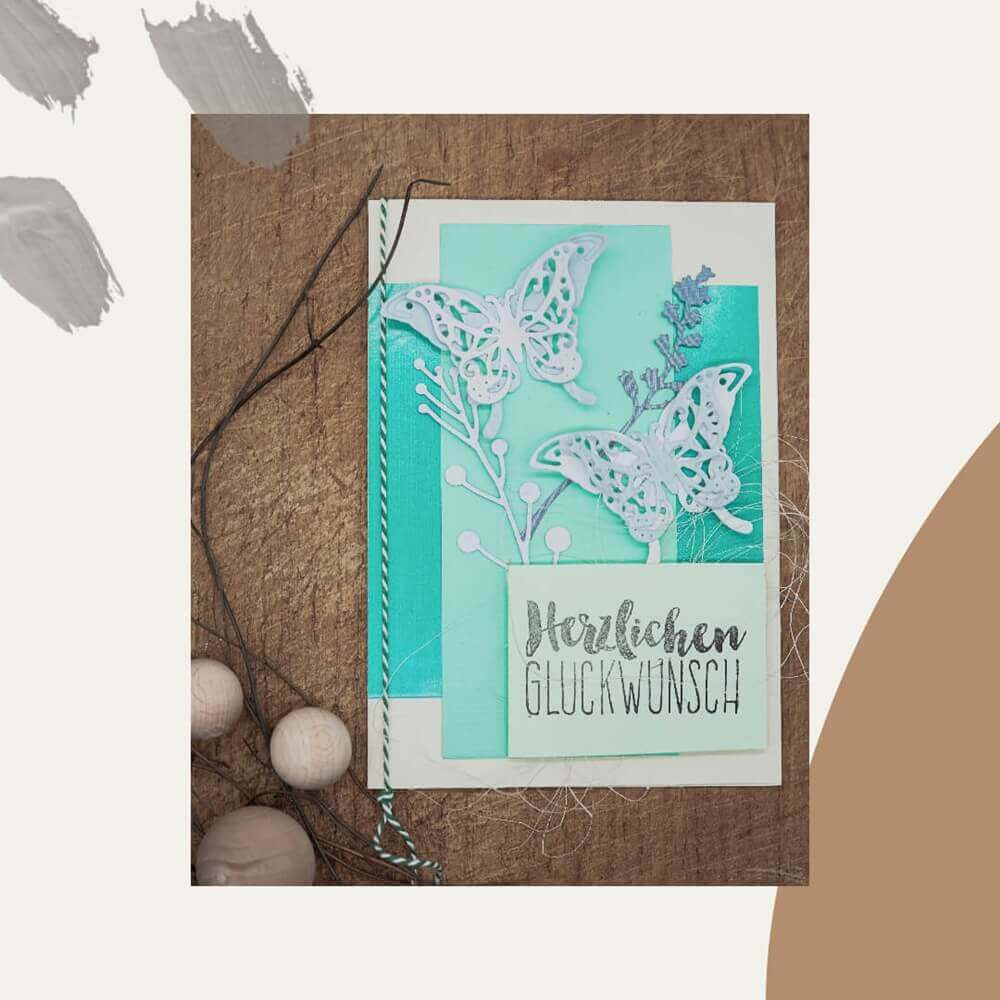Eine Grußkarte mit geschichtetem blauem und weißem Papier, Stanzschablone: Zwei Schmetterlinge von Stanzenshop.de, dekorative Schmetterlinge und "Herzlichen Glückwunsch"-perfekt zum Basteln oder Scrapbooking auf einer Holzunterlage mit Schnur und Holzperlen.