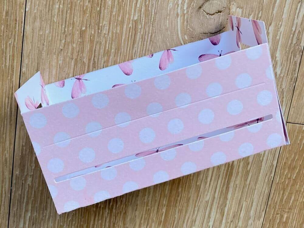 Eine rosafarbene rechteckige Pappschachtel mit weißen Tupfen und Schmetterlingsmuster im Inneren, hergestellt mit der Stanzschablone: Großer Karton von Stanzenshop.de, perfekt als Geschenkbox Hochzeit und für Bastelprojekte, abgebildet auf einer Holzunterlage.