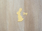 Ein gelber Papierausschnitt eines sitzenden Hasen mit einer Schleife, hergestellt mit der "Stanzschablone: Hase mit einer Fliege" von Stanzenshop.de, wird auf hellem Holz präsentiert - eine perfekte Idee fürs Basteln zu Ostern.