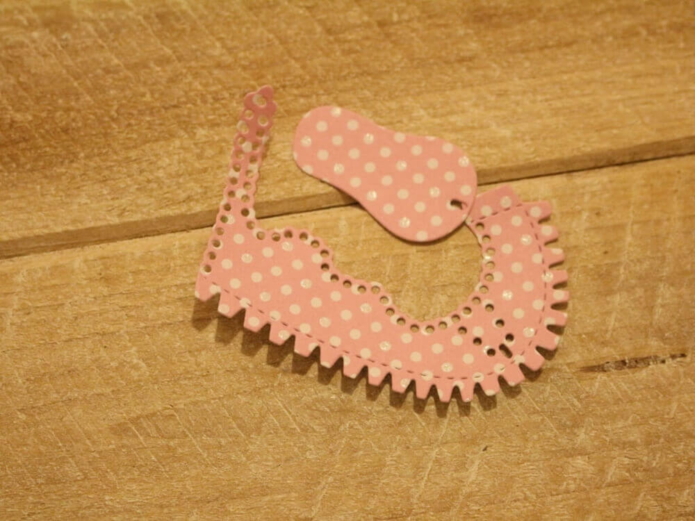 Zwei Stücke rosafarbenes, gepunktetes Papier, zugeschnitten mit der "Stanzschablone Kinderschuh für ein Baby" von Stanzenshop.de, werden dekorativ geformt und auf eine Holzunterlage gelegt.