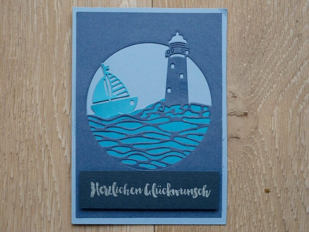 Die Stanzschablone Leuchtturm und Meer in rundem Rahmen von Stanzenshop.de schneidet eine blau-weiße Segelboot- und Leuchtturmszene mit "Herzlichen Glückwunsch" aus, perfekt für Basteln oder Scrapbooking-Projekte.