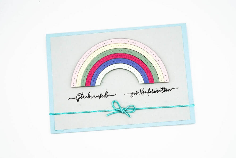 Eine Grußkarte mit einem geschichteten Regenbogendesign mit der Stanzschablone Regenbogen von Stanzenshop.de, handgeschriebenem deutschen Text und einer blauen Schleife - ein inspirierendes Bastelergebnis, kompatibel mit gängigen Stanzmaschinen.