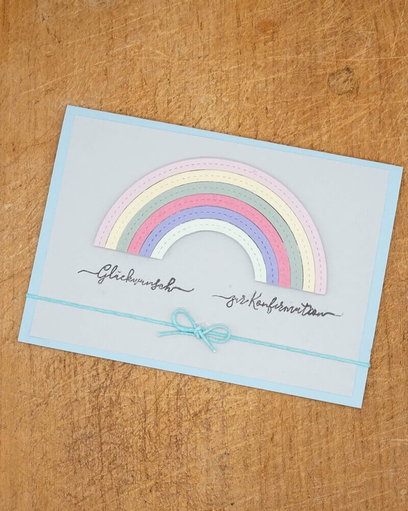 Die pastellfarbene Regenbogenkarte aus der Stanzschablone Regenbogen von Stanzenshop.de mit kursiver Schrift und blauer Schnurschleife ruht auf Holz - perfekt, um Ihr Bastelergebnis zu präsentieren.