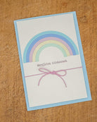 Eine Grußkarte mit einem pastellfarbenen Regenbogen aus der Stanzschablone Regenbogen von Stanzenshop.de, mit "Herzlichen Glückwunsch" auf Deutsch und einem rosafarbenen Streichbogen, ausgestellt auf Holz für ein inspirierendes Bastelergebnis.