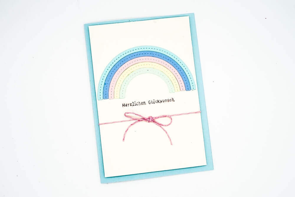 Eine Grußkarte mit der Stanzschablone Regenbogen von Stanzenshop.de zeigt ein pastellfarbenes Regenbogendesign, den Schriftzug "Herzlichen Glückwunsch" und eine rosa Schleife auf hellblauem Hintergrund.
