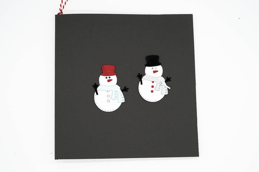 Eine schwarze Karte mit zwei Schneemännern - einer mit roter Mütze und Schal, einer mit schwarzer Mütze und weißem Schal - hergestellt mit der "Stanzschablone: Schneemann mit Schal und Zylinder" von Stanzenshop.de, perfekt für winterliche Bastel- oder weihnachtliche Stanzprojekte.