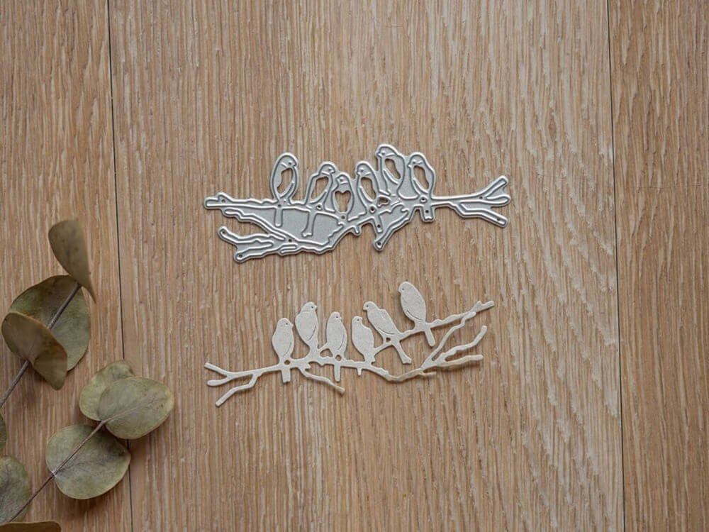 Die Stanzschablone "Sechs Vögel auf einem Ast" von Stanzenshop.de zeigt sechs Vögel auf einem Ast, dargestellt auf Holz mit getrockneten Blättern - ideal für Kartenherstellung oder Scrapbooking.
