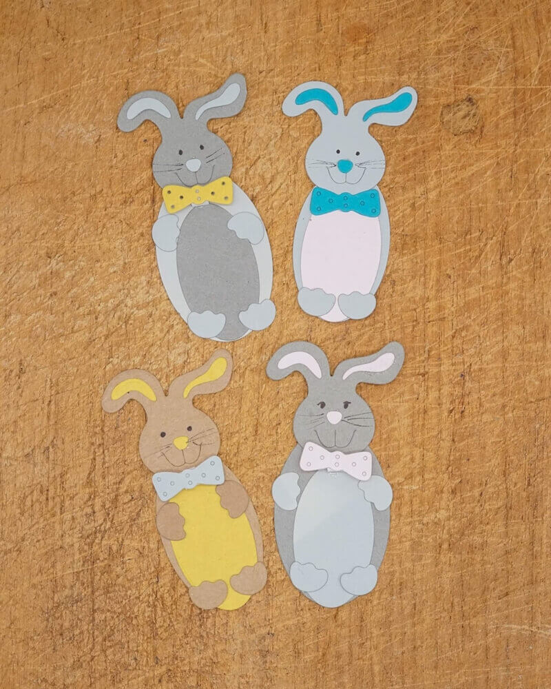 Vier Papier-Osterhasen mit Fliege in bunten Farben, arrangiert auf Holz - ideal zum Basteln zu Ostern mit der Stanzschablone "Süßer Osterhase" von Stanzenshop.de.