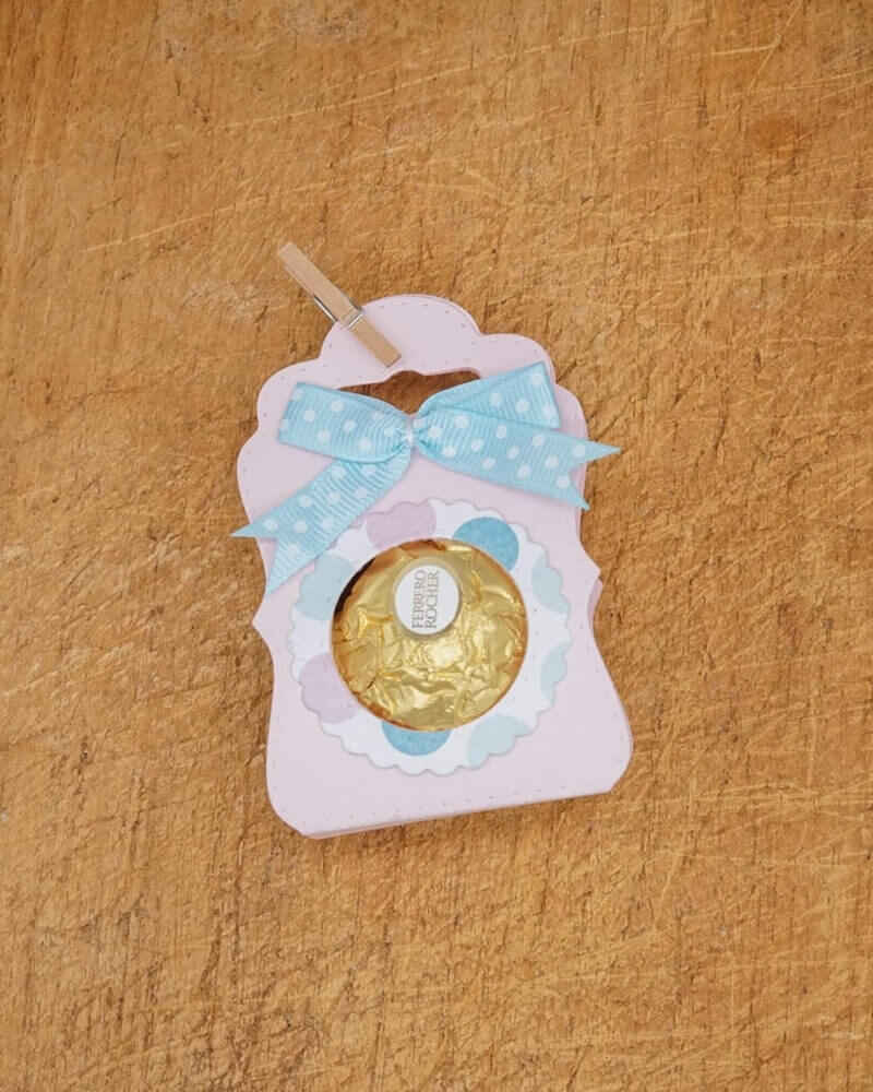 Ein Ferrero Rocher auf einer pastellrosa und blauen Karte, hergestellt mit der Stanzschablone Tischkarte mit Kreis von Stanzenshop.de, verziert mit einer blauen Tupfenschleife - ideal für Scrapbooking oder Bastelset-Projekte.