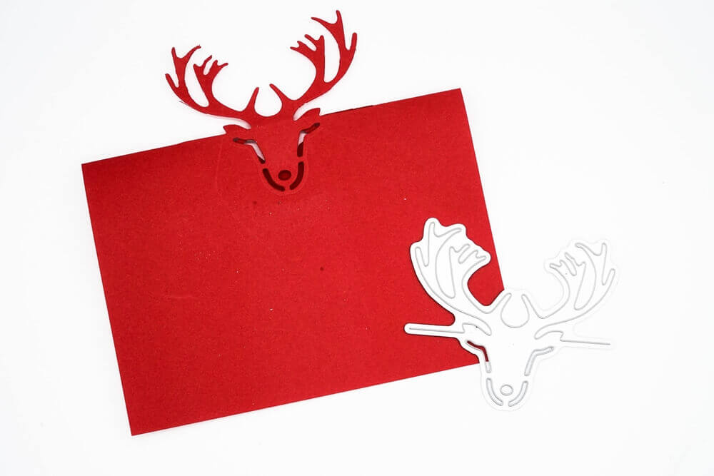 Eine rote Karte mit einem ausgeschnittenen Rentierkopf aus der Stanzschablone: Die Tischkarte Rentierkopf von Stanzenshop.de liegt neben einer weißen Rentierschablone aus Metall - ideal für Tischkarte Basteln oder Scrapbooking Schablonen Projekte.