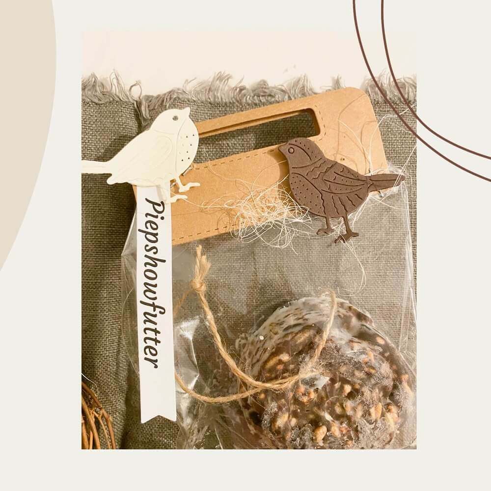 Eine verpackte Vogelfutterkugel mit einem "Piepshowfutter"-Anhänger, dekoriert mit der Stanzschablone Vier verschiedene Vögel von Stanzenshop.de in Weiß und Braun, ausgestellt auf strukturiertem Stoff.