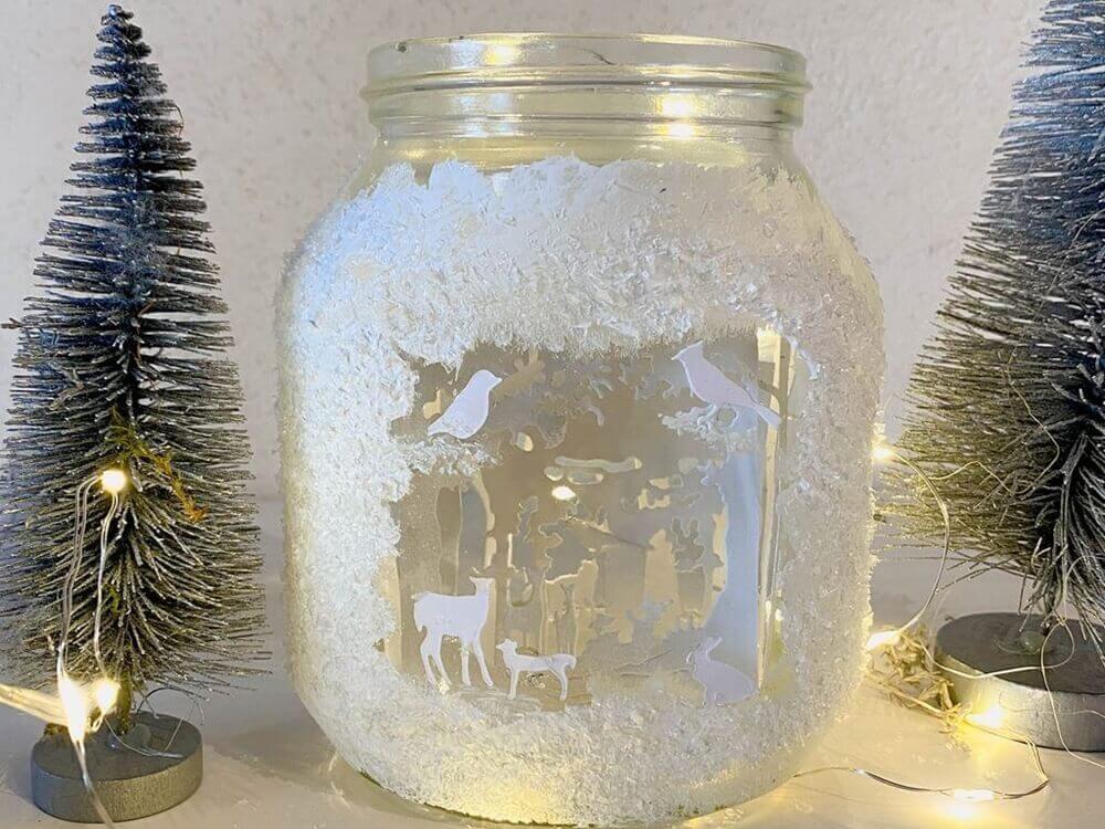 Ein dekoratives Winterglas mit einer ausgeschnittenen Waldszene, umgeben von einer Stanzschablone Wald mit Tieren und Lichterketten von Stanzenshop.de.