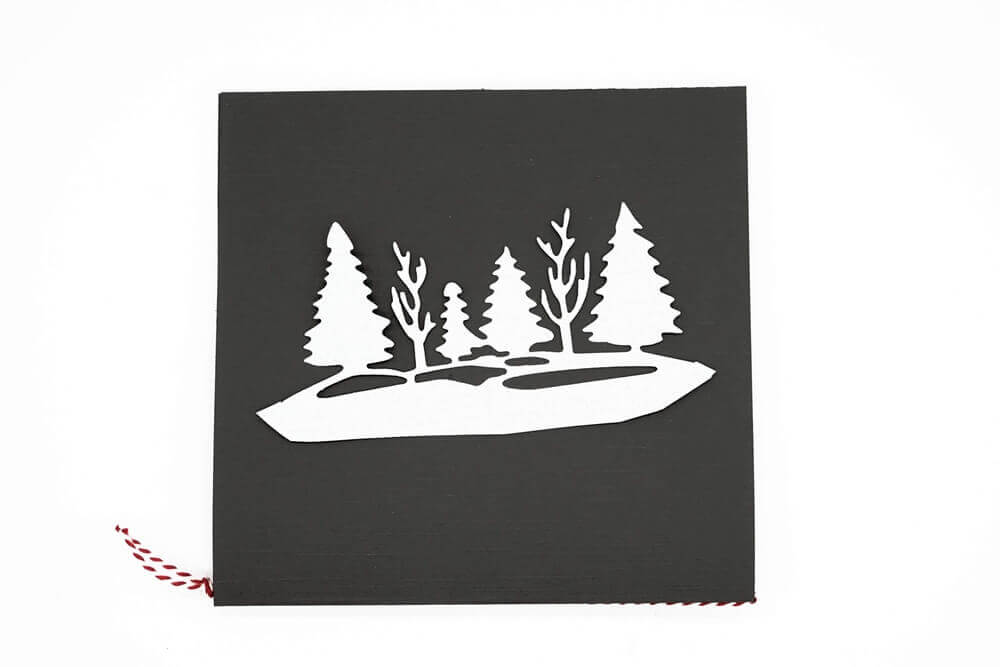 Eine schwarze quadratische Karte zeigt einen weißen Ausschnitt von Tannen und kahlen Bäumen, hergestellt mit der "Stanzschablone: Waldlandschaft im Winter" von Stanzenshop.de. Eine rot-weiße Kordel akzentuiert den linken Rand und sorgt für festlichen Charme.